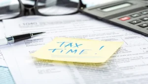 Tax forms with calculator, glasses, pen and yellow sticky note reading 'TAX TIME!' on 1040 form