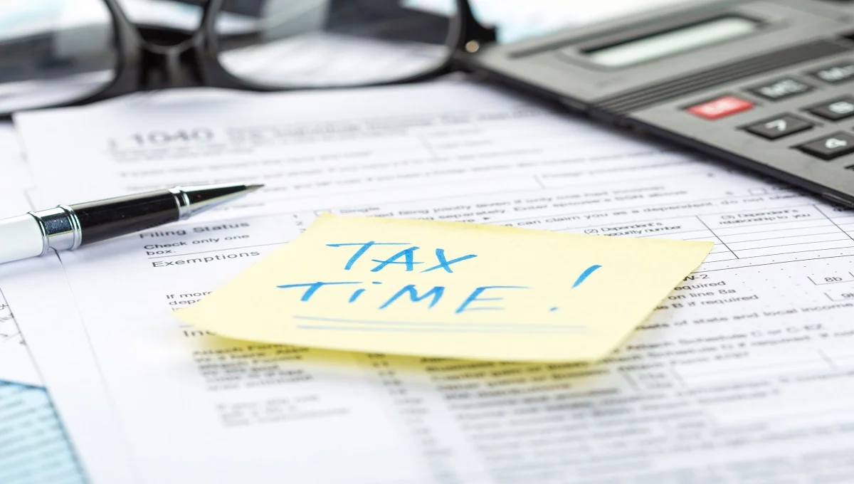 Tax forms with calculator, glasses, pen and yellow sticky note reading 'TAX TIME!' on 1040 form